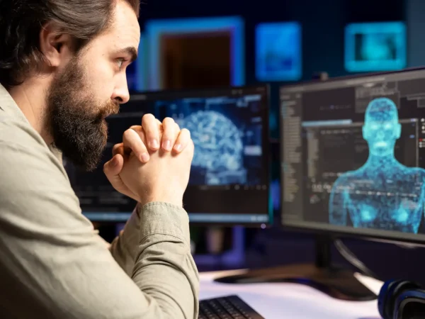Homem concentrado em frente a dois monitores com imagens médicas e um modelo 3D de corpo humano, em ambiente de trabalho com tecnologia e software, voltado à integração entre sistemas digitais e operações presenciais, tema de omnicanalidade