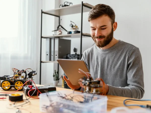 Técnico em laboratório industrial analisa anotações e controla componentes eletrônicos em bancada, enquanto um pequeno robô montado ao lado indica prática de recrutamento e treinamento de operadores em escassez de mão de obra técnica.
